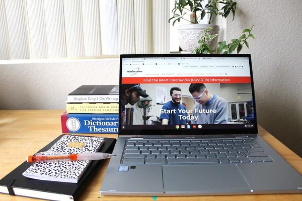 An open laptop showing a COVID-19 information webpage on Palomar College's website. Three books rest behind it and a notebook and mechanical pencil rest to the left.