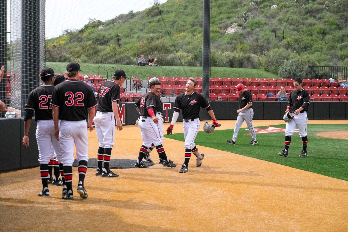 A male Palomar baseball player walks to the left with his helmet off as several teammates congratulate him from the left.