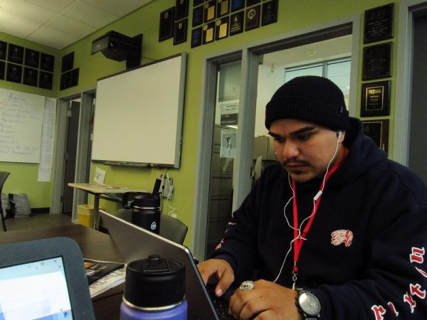 A male journalism student types on his laptop, sporting a pair of white earbuds