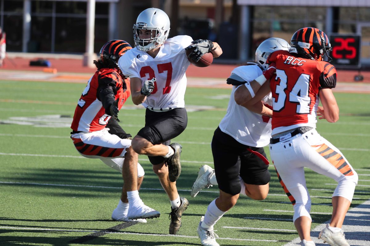 A Palomar football player (37) runs pass an opposing player, carrying a football in his left arm. A team player and an opposing player tackle each other on the right.