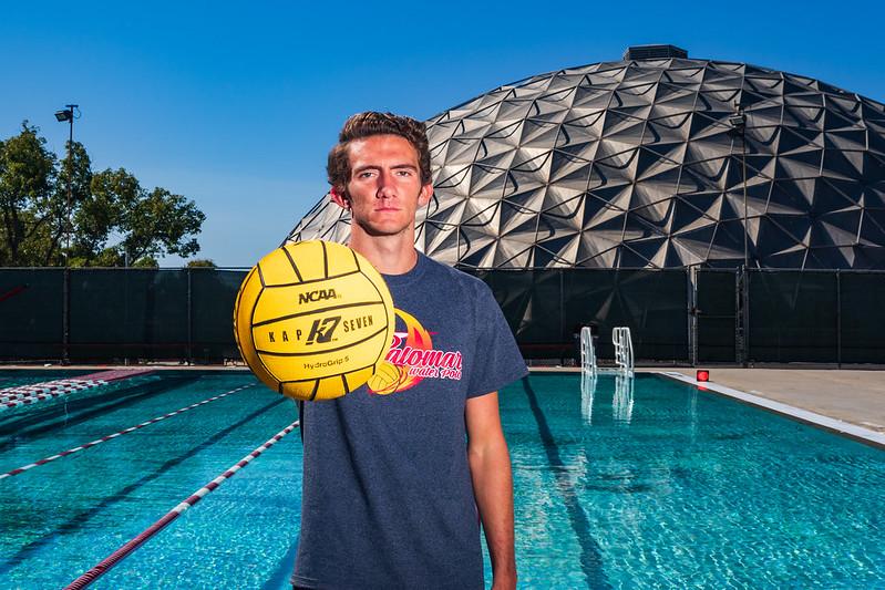 Palomar water polo player Kevin McCollum stands and holds a yellow ball in front of him toward the view. A swimming pool and The Dome are behind him.