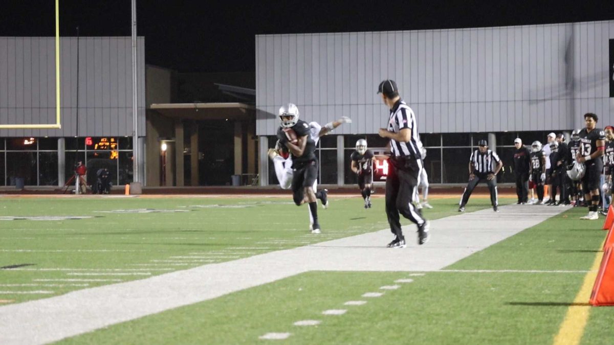 A football player in a black uniform runs across a field near the border while a referee jogs in front of him and looks back. Several players are standing on the sidelnes.