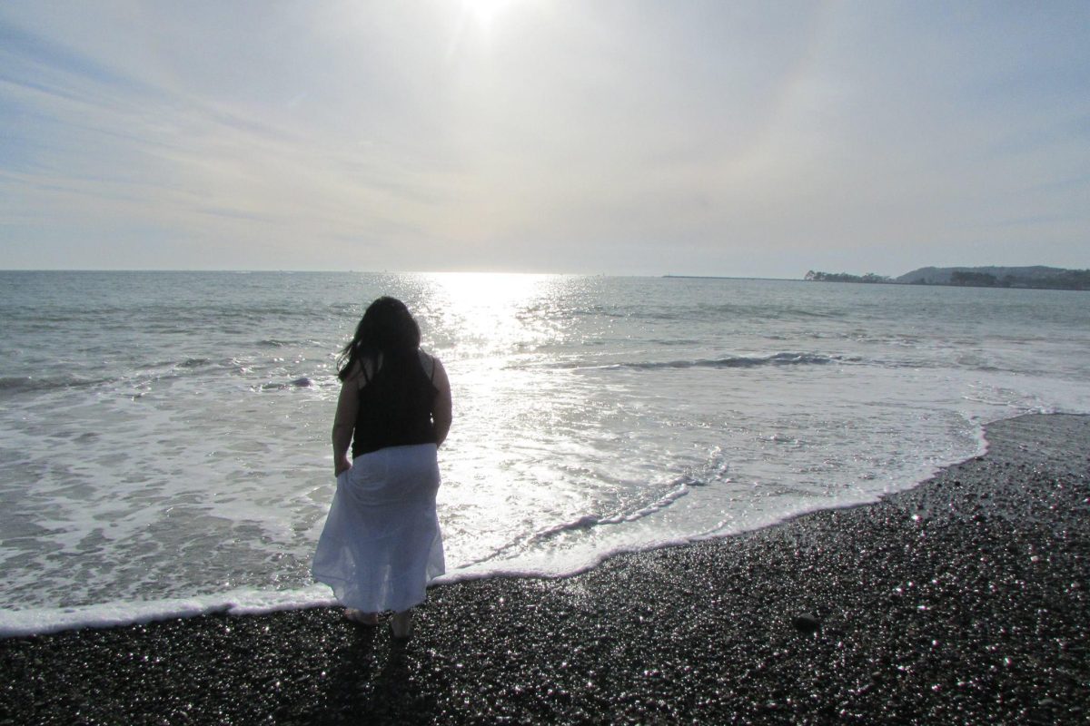 Woman with long, black hair watches waves splash on the shore on Capistrano Beach in Orange County. She stands on the shore riddled with round, smooth pebbles.