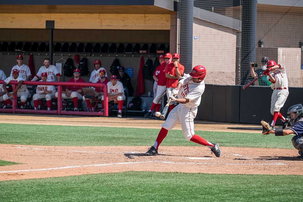 A male Palomar baseball player in a white jersey and pants swings a baseball bat with the baseball inches from the bat. A dozen team players watch from the dugout in the background.