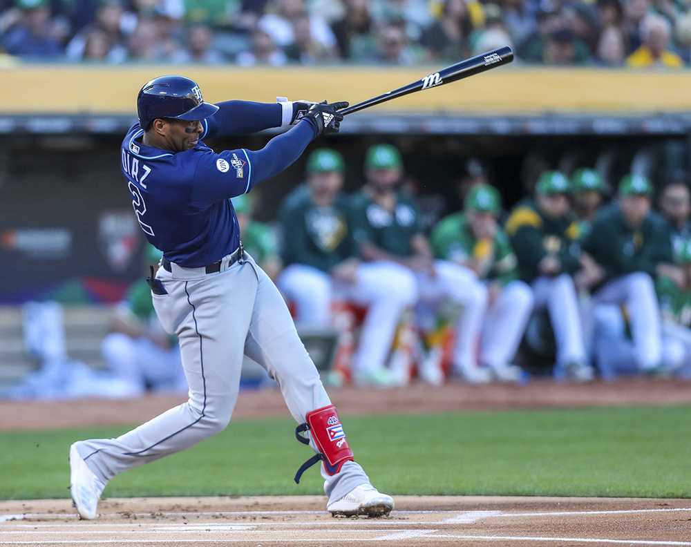 Tampa Bay Rays’ Yandy Diaz swings a baseball bat, hitting a home run. Several players from the opposing team watch and sit at the dugout.