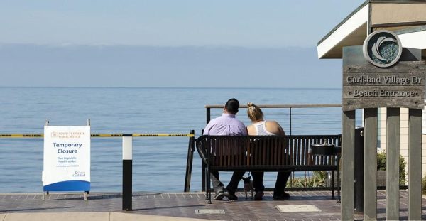 A couple sits on a bench near a closure sign at a Carlsbad beach.