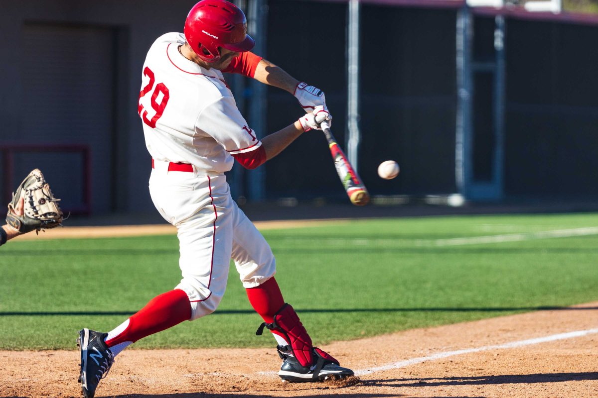 A male Palomar baseball player in a white jersey and pants swings a baseball bat with the baseball inches from the bat.