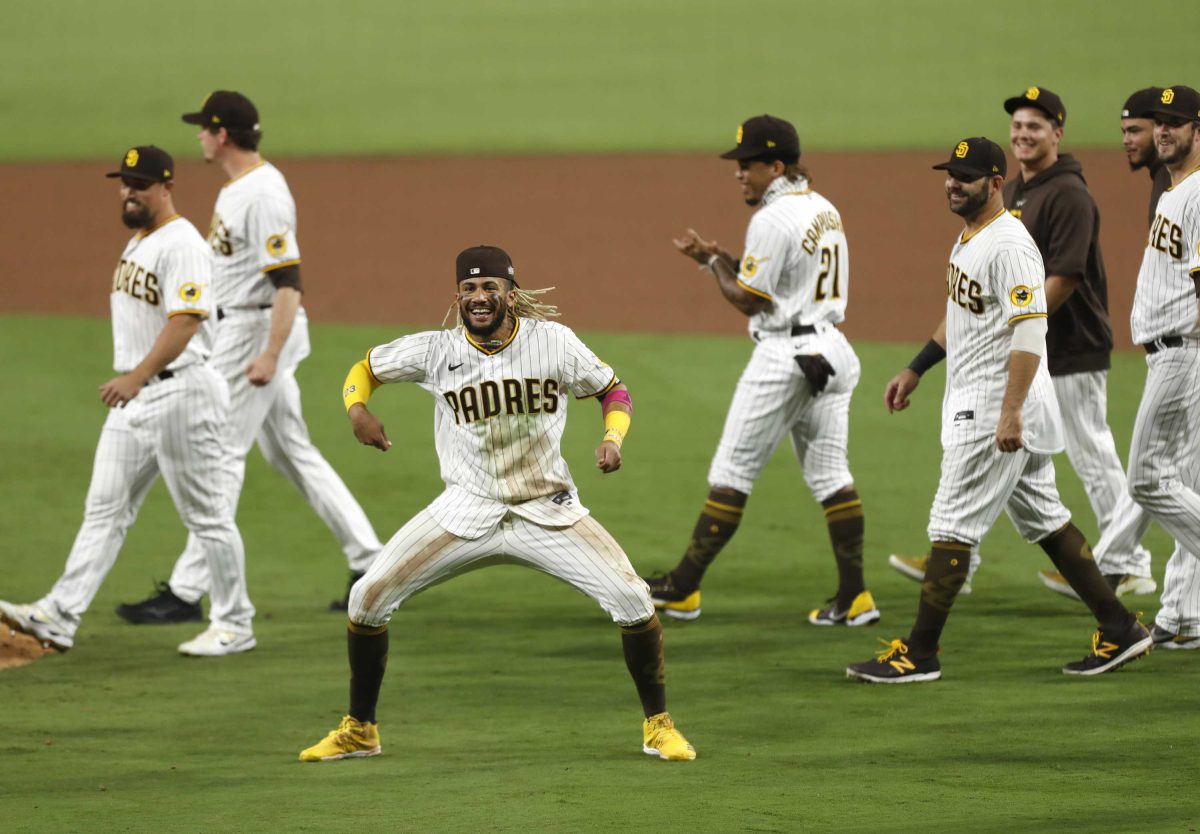 A San Diego Padres baseball player dances with his legs in a wide squat stance and arms bent to his sides. A group of his team players walk to the left behind him.