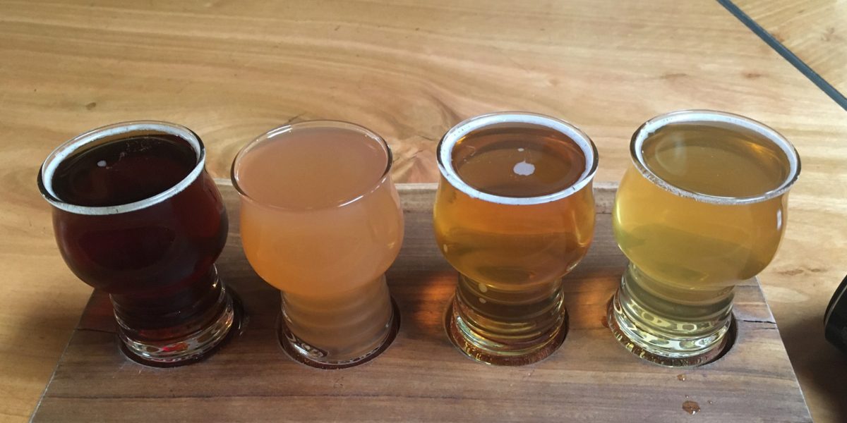 A flight of four apple ciders on a wooden tray on a wooden bar.