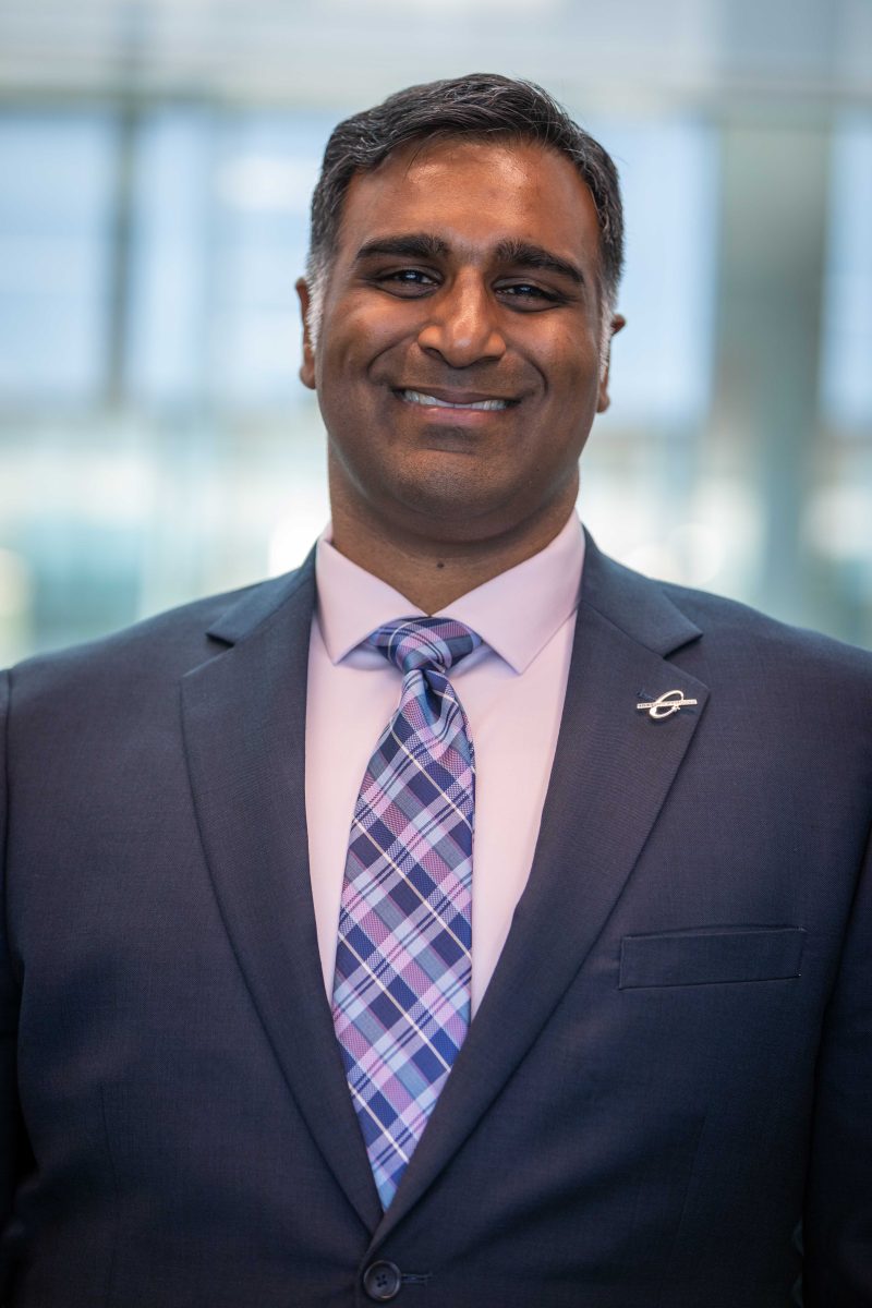 Portrait of a man of Indian descent smiling, wearing a dark grey suit and a checkered tie.