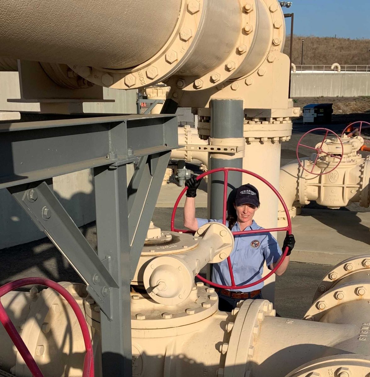 A woman in a blue shirt turns a large, red, metal wheel with both hands. Large beige-colored pipes are above, behind, and in front of her.