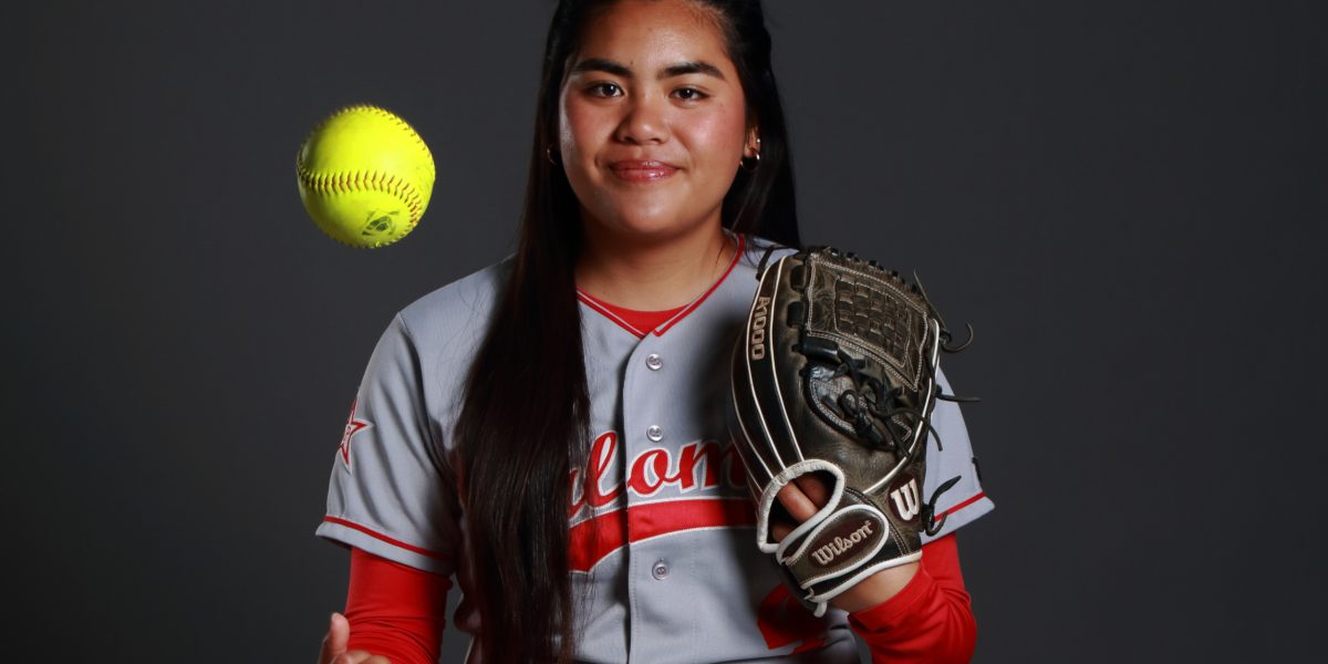 Palomar softball player Alyssa Domingo wears her gray and red uniform and catcher's mitt while tossing a lime green softball in the air.