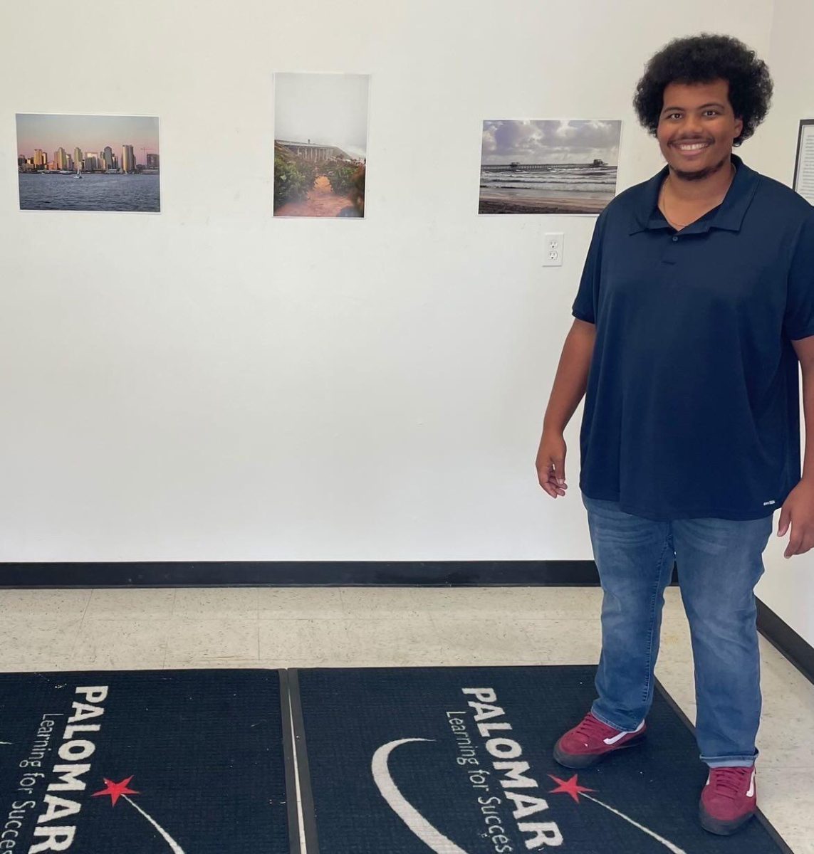 Dominic Brown stands in front of his artwork. Three photos hang on the wall behind him.