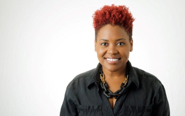 Portrait of Cassandra Schaeg smiling and sporting red, curly hair, a dark gray shirt with two breast pockets,and a black and gold necklace that resembles a linked chain.