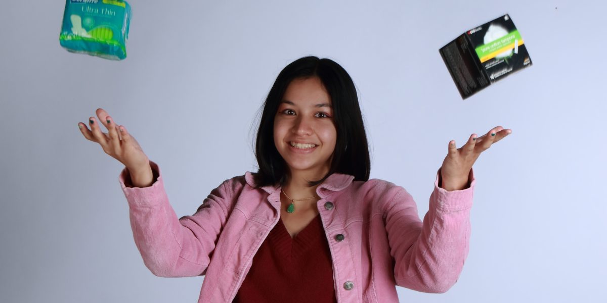 Palomar student Gabby Le tosses up a pack of tampons on each hand with a grin on her face. She wear s a pink courduroy jacket and a red top.