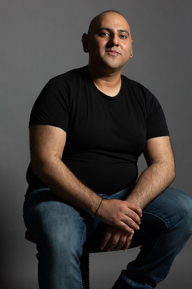 Zeshaun Hassan sits on a stool in a relaxed pose with a gray background.