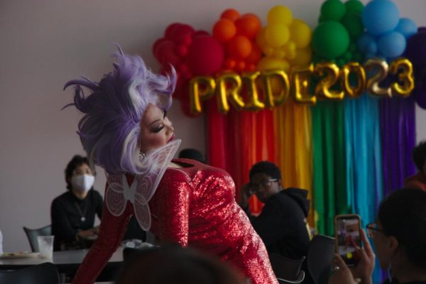 drag queen in a glittery red dress and purple wig performing expressively in front of a wall of rainbow balloons at a college event