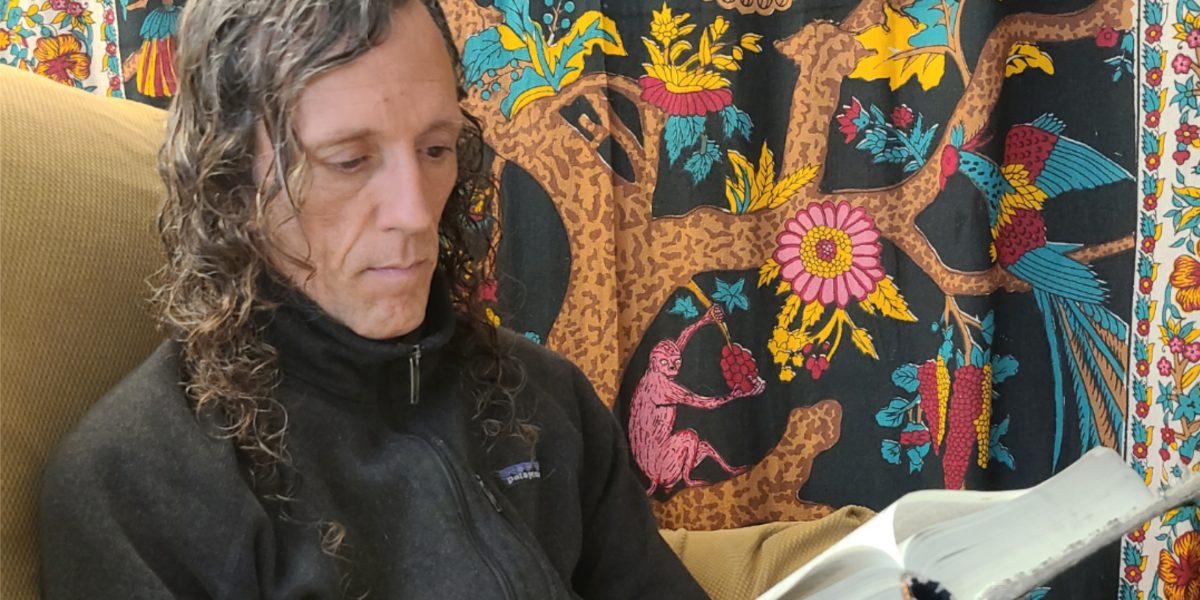 man with longm, wavy brown and gray hair sits on a couch and reads a big book with a colorful tapestry in the background.