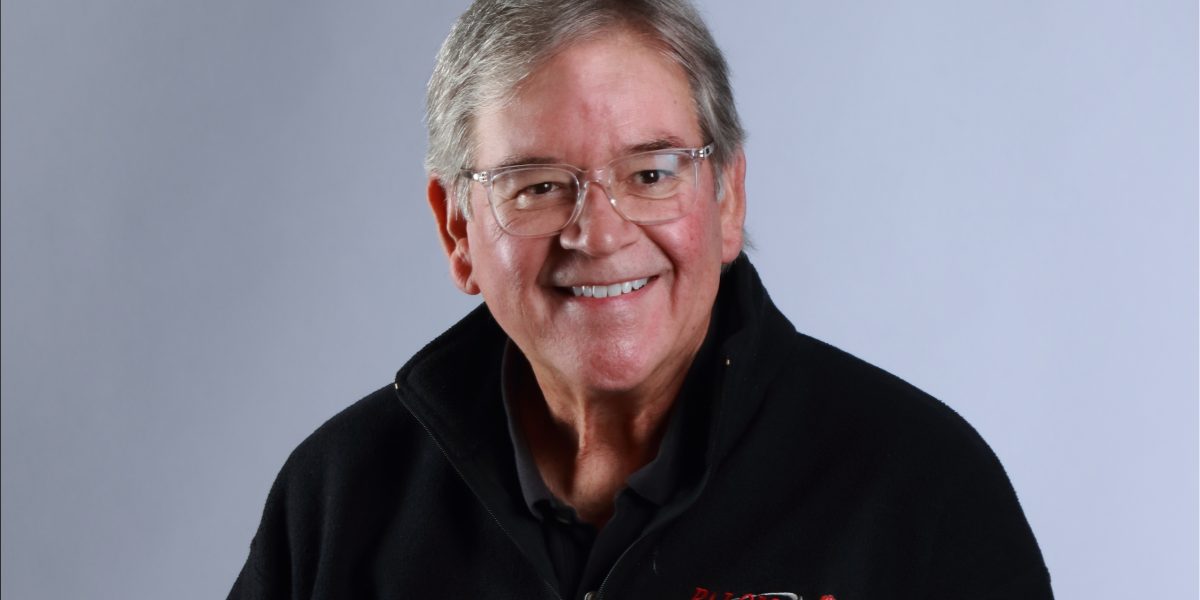 Governing board member Mark Evilsizer smiles and sits on a stool with his hands folded. He wears light blue jeans and a black light jacket with a Palomar College logo.