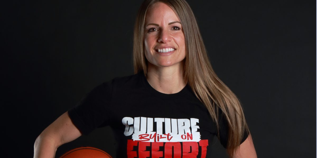 Head basketball coach Leigh Marshall smiles and stand with her left hand on her hip and holds a basketball to against her hip with her right hand. She wears a black T-shirt that says "Culture built on effect."