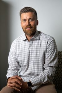 Dr. Patrick Savaiano sits with his hands folded, wearing a gray and white grid-pattern shirt and brown trousers.