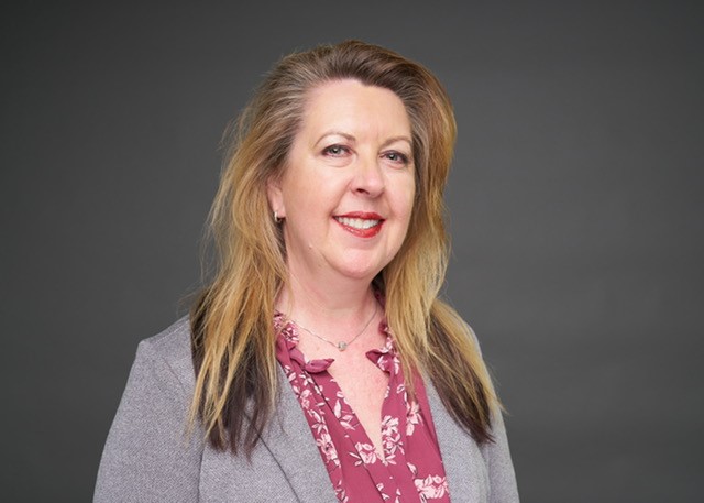 Dr. Tina Recalde in a grey suit and magenta shirt with white flower patterns.