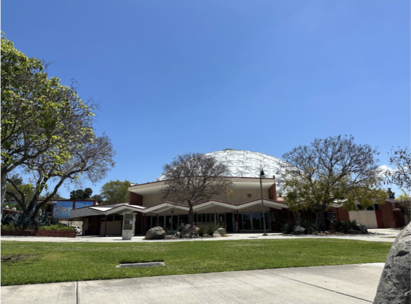 Palomar's dome-shaped gray building sites in the background. In the foreground is grass and a sidewalk.