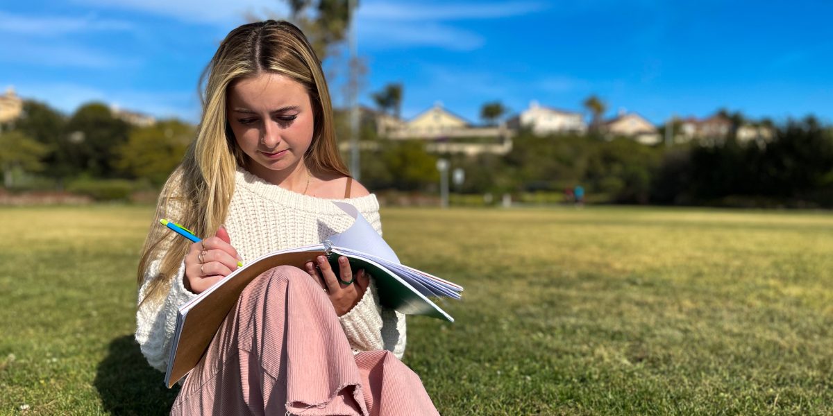 Female student sits on a grassy park writing in her notebook on a sunny day.