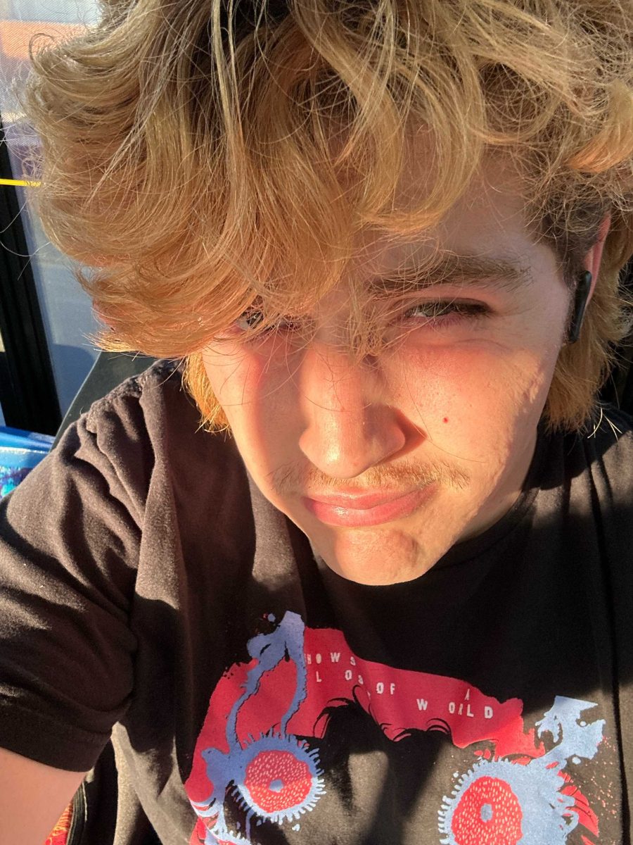 A man with blonde, curly hair is making a face and in partial shadow. He wears a black shirt.