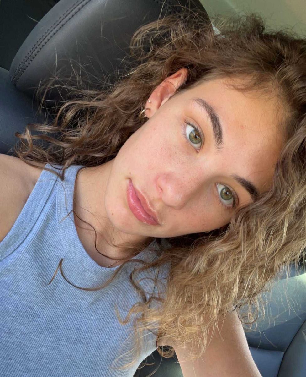 A young woman with curly dark blonde hair is staring beyond the camera and not smiling.