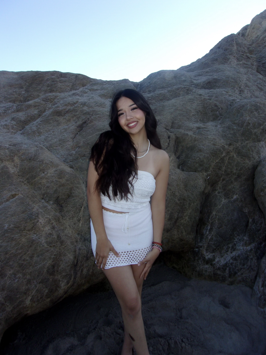 A brunette with curly hair over one should poses outside in a white dress. She is smiling.