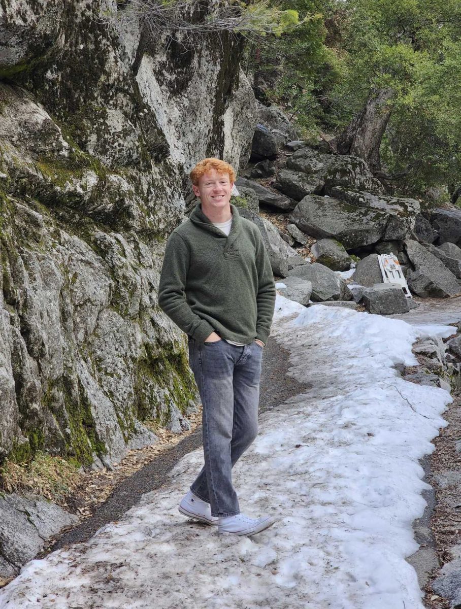 Red haired male stands in the snow outside. He wears a green sweater and jeans. He is smiling.