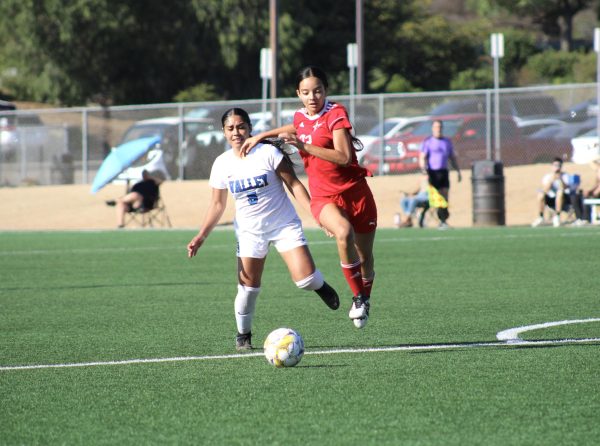 Palomar soccer player in a red jersey with number 32 in white on it runs on a green soccer field to reach a white and yellow soccer ball before San Bernardino soccer player with a white jersey with a blue number 5 is running behind trying to catch up.