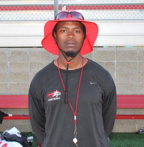 A Palomar football coach in a red bucket hat and long-sleeve black shirt with a Palomar football logo poses for a photo.