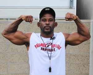 A Palomar football coach in a black and white hat and a white tank-top with a Palomar football logo flexes his arms while posing for a photo.