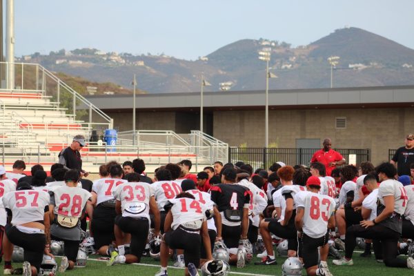 Three Palomar football coaches stand on a football field while a team of football players kneel in front of the coaches.