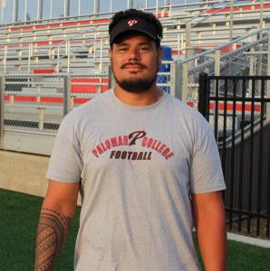 A Palomar football coach with a tattoo on his right forearm wears a black visor and a grey shirt both with a Palomar football logo poses for a photo.