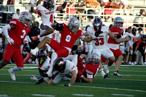 Palomar player number 6 in a red uniform falls over a diving Ventura player in white attempting to dodge and surrounded by other players from both teams.