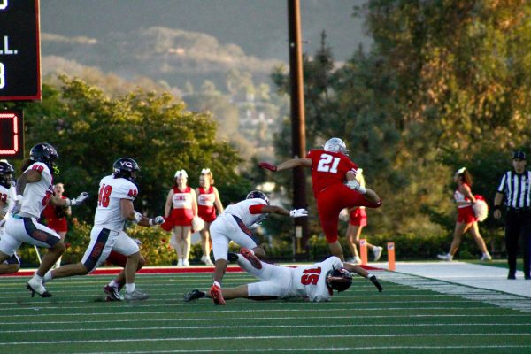 Palomar player in a red uniform number 21 jumps over a diving Ventura player in a white uniform number 15 as he runs up the football field.