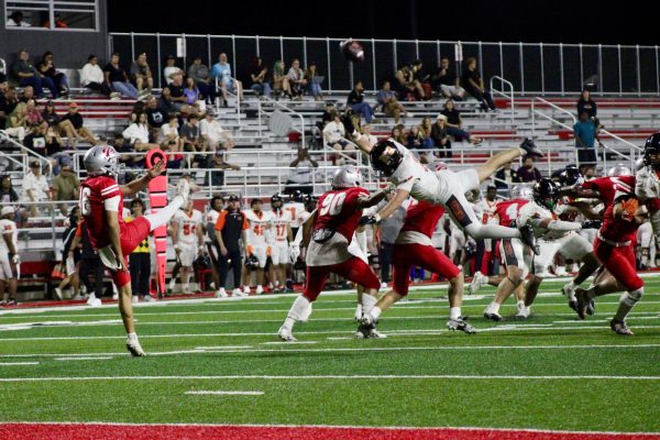 Palomar's kicker in red is kicking the football while one of Ventura's players dives towards the kicker trying to block the ball.