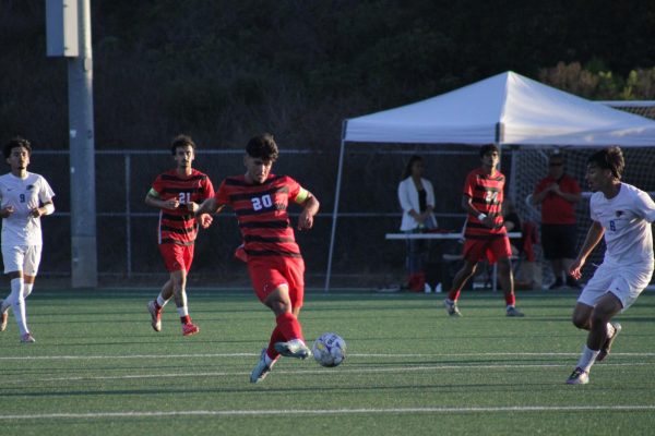 Sam Cobain (#20) is in the center with his left arm in front of him and his right arm behind him and his right leg his out in front of him as he passes the white and yellow soccer ball to a teammate nearby.