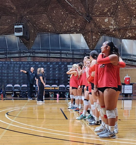 Palomar volleyball players in red jerseys line up at the back of the volleyball courts with their coaches in black watching on before a game.