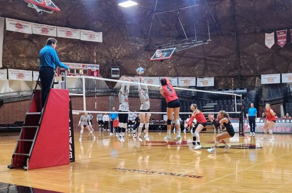 Palomar player in a red jersey, number 9 hits a volleyball over the outstretched arms of two Santa Ana players in white jerseys.