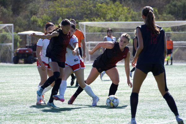 Cisilya Martinez (#7) is wearing a black and red uniform and is kicking the ball away from the opposing player, who has her leg out in front of her to try to take the ball away from Martinez. Palomar's Naomi Allen (#24) is also wearing a black and red uniform, looking towards Martinez, ready to assist as needed.