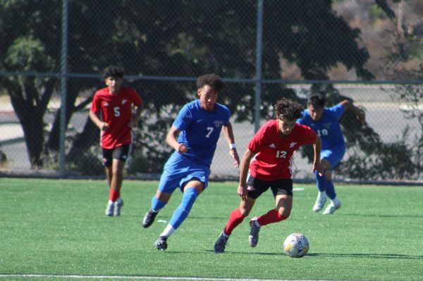 Palomar player number 12 in red dribbles a soccer ball as he moves away from a pursuing Santa Monica player number 7 in blue.