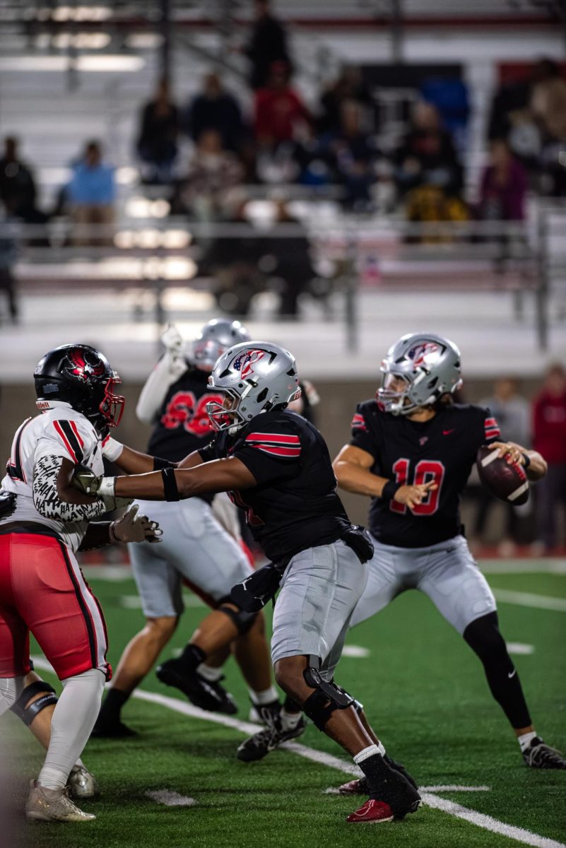 Palomar quarterback Jakob Harris (#10) in black looks down field and prepares to pass the football while his offensive linemen block Mt San Jacinto players in white from tackling him.