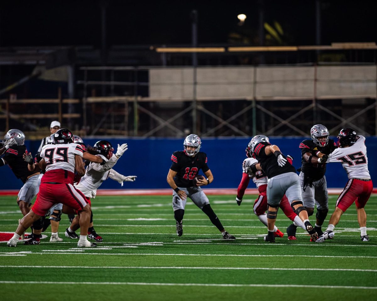Palomar quarterback Jakob Harris #10 in black holds the ball in his left hand as he moves to run up field while players on Mt San Jacinto in white attempt to tackle him.