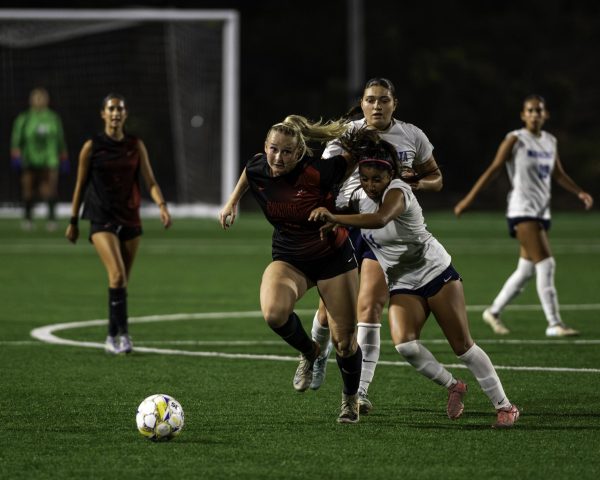 Palomar player in black is attempting to push herself away from a MiraCosta player in white as both try to run to gain possession of the soccer ball near the midfield.