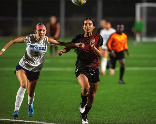 Palomar player in black races alongside a MiraCosta player in white to get possession of the soccer ball near MiraCosta's goal.