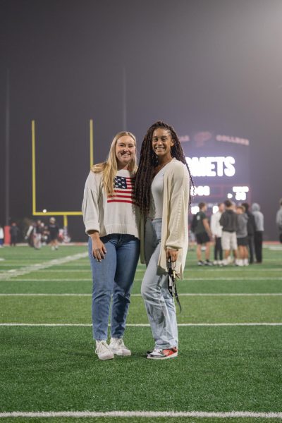 Cara Heise wears a white sweater with the American flag on it and stands next to Bianca Littleton on a football field with the scoreboard in the background.